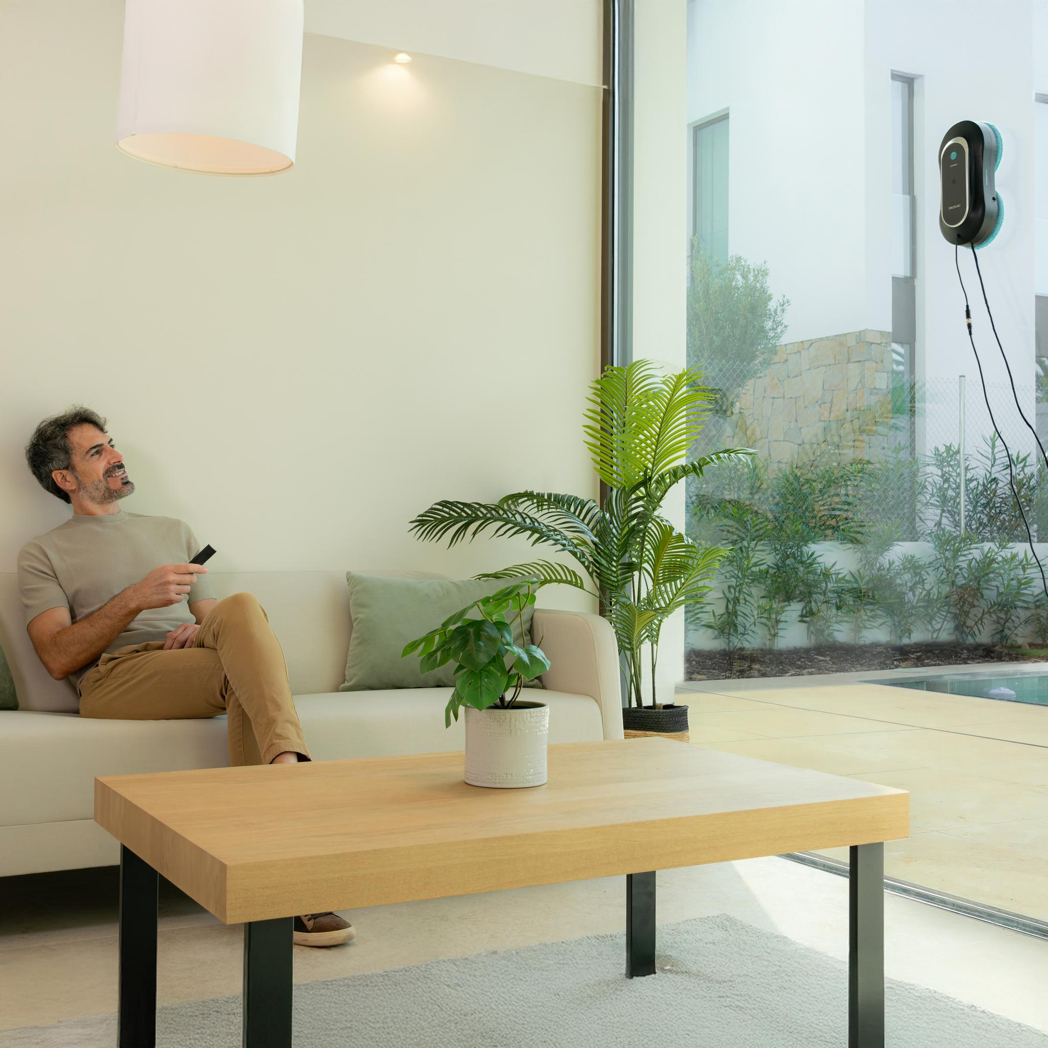 Hombre en sofá usa mando. Limpiador de ventanas limpia. Plantas y mesa en la habitación.