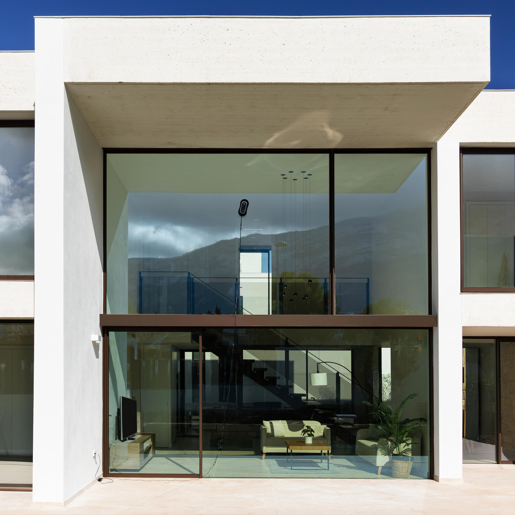 Edificio blanco moderno con grandes ventanales. El interior incluye sofá, mesa y escaleras. Cielo azul visible.