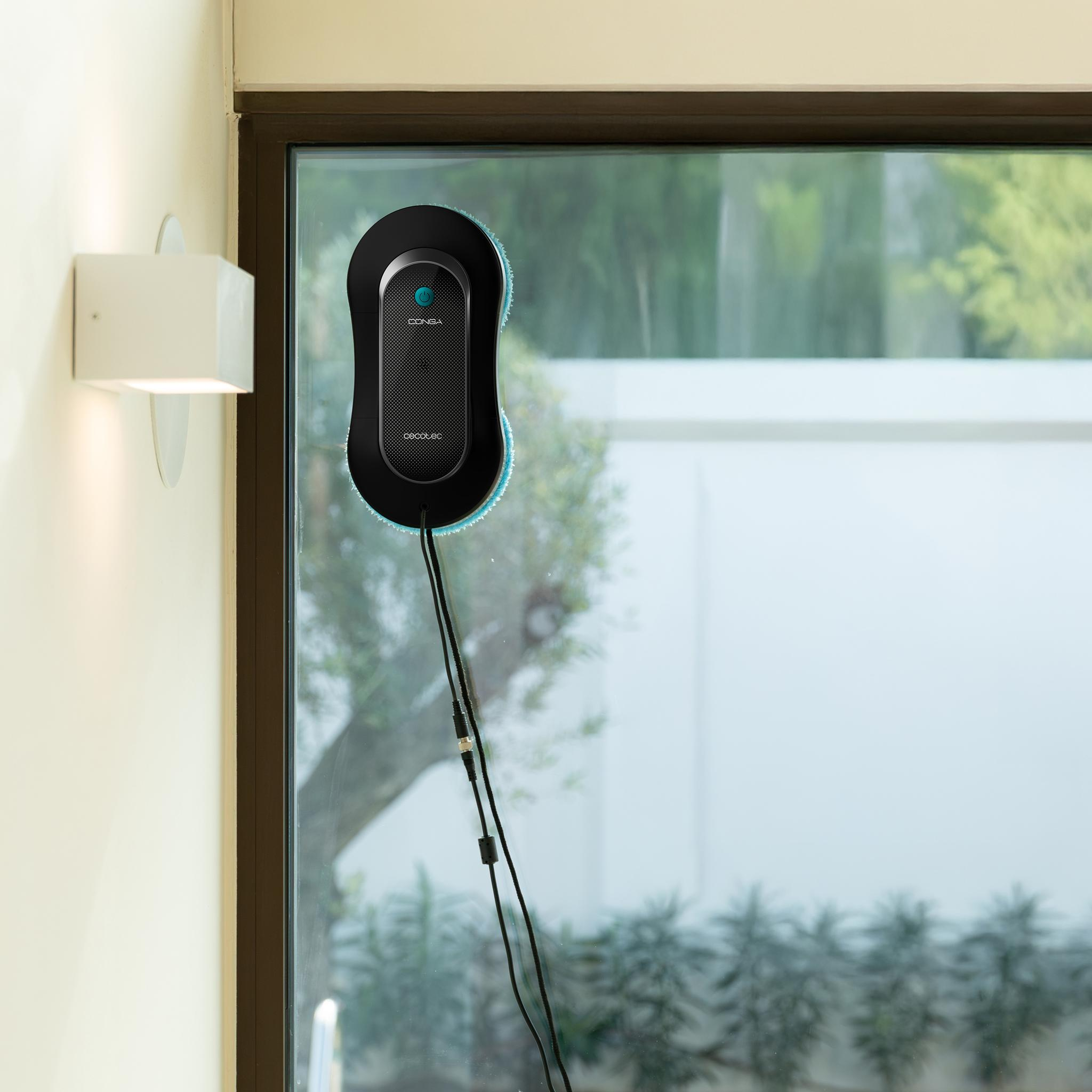Un robot limpiador de ventanas negro en un cristal. Una lámpara blanca en la pared. Afuera, se ven árboles verdes.