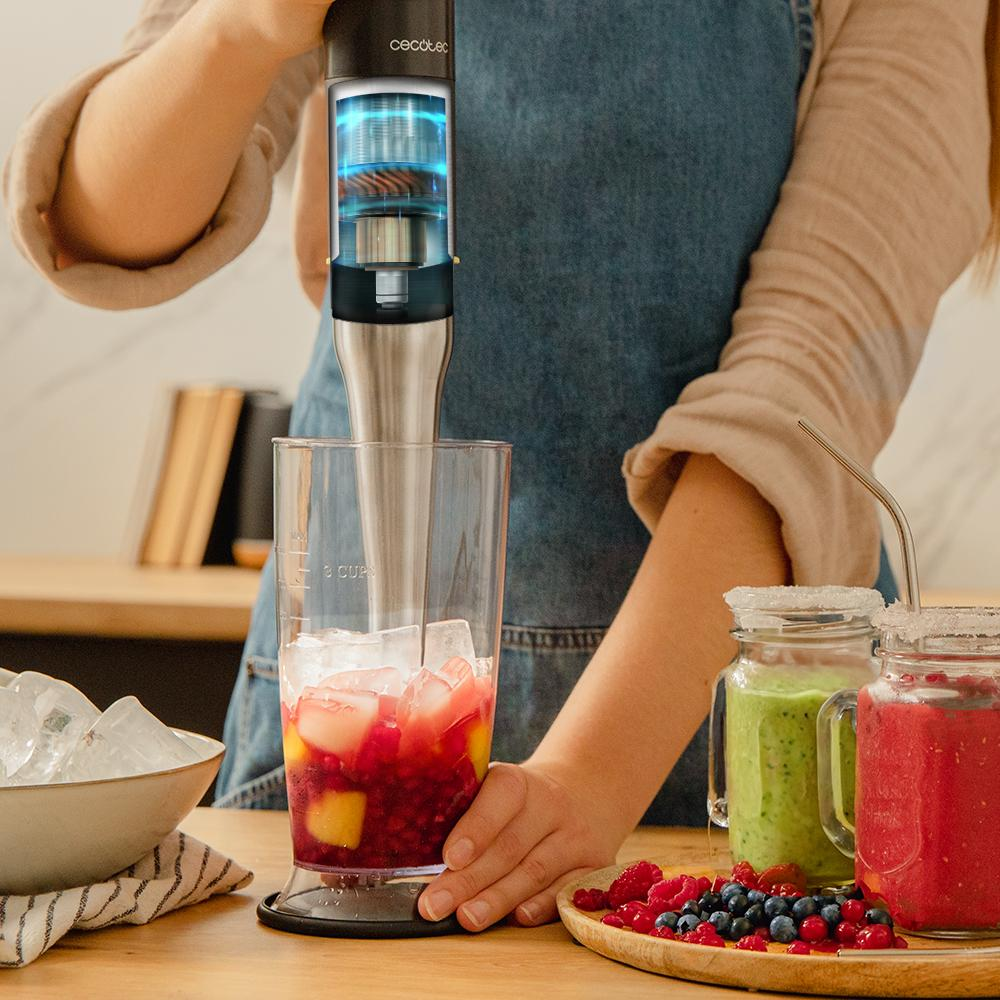 Una persona mezcla una bebida con una batidora de mano. El vaso contiene hielo y fruta. Frascos de batidos y plato de bayas cerca.