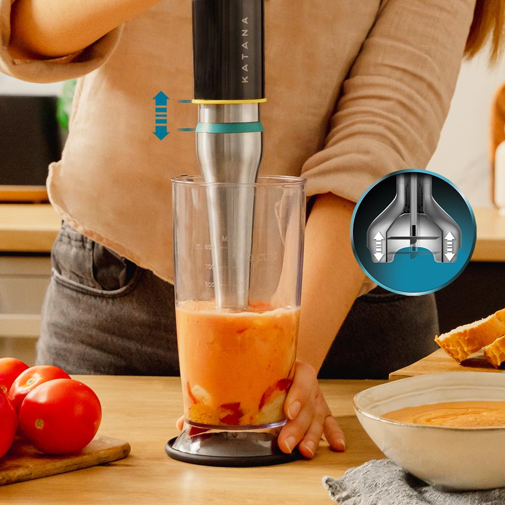 Una persona usa una licuadora en un vaso para mezclar sopa naranja. Tomates, un cuenco de sopa y rebanadas de pan están sobre la mesa.