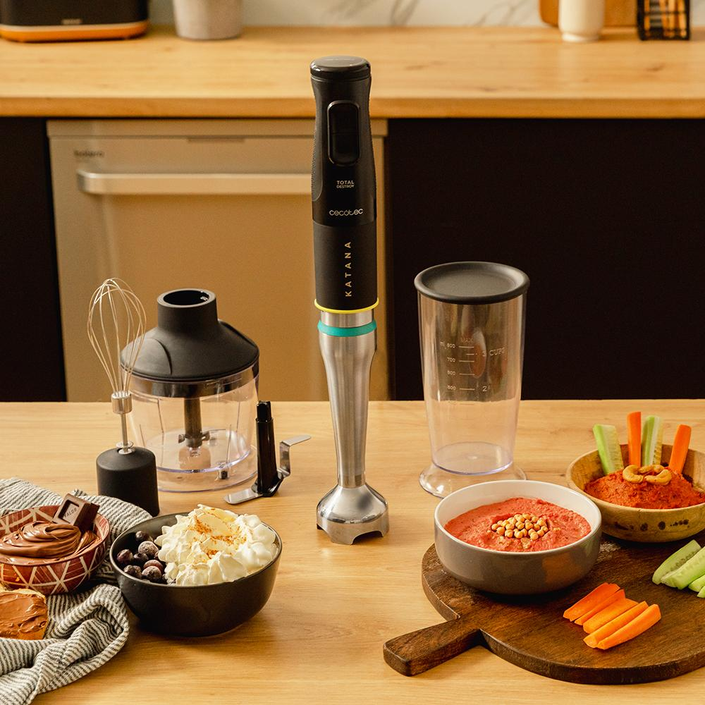 Escena de cocina con batidora, comida e ingredientes sobre una mesa de madera. Incluye cuencos con hummus, crema y verduras.