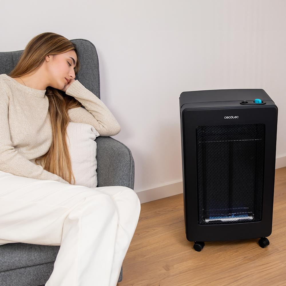 Una mujer duerme en una silla gris junto a un calefactor negro. Viste suéter beige y pantalones blancos, con la cabeza apoyada.