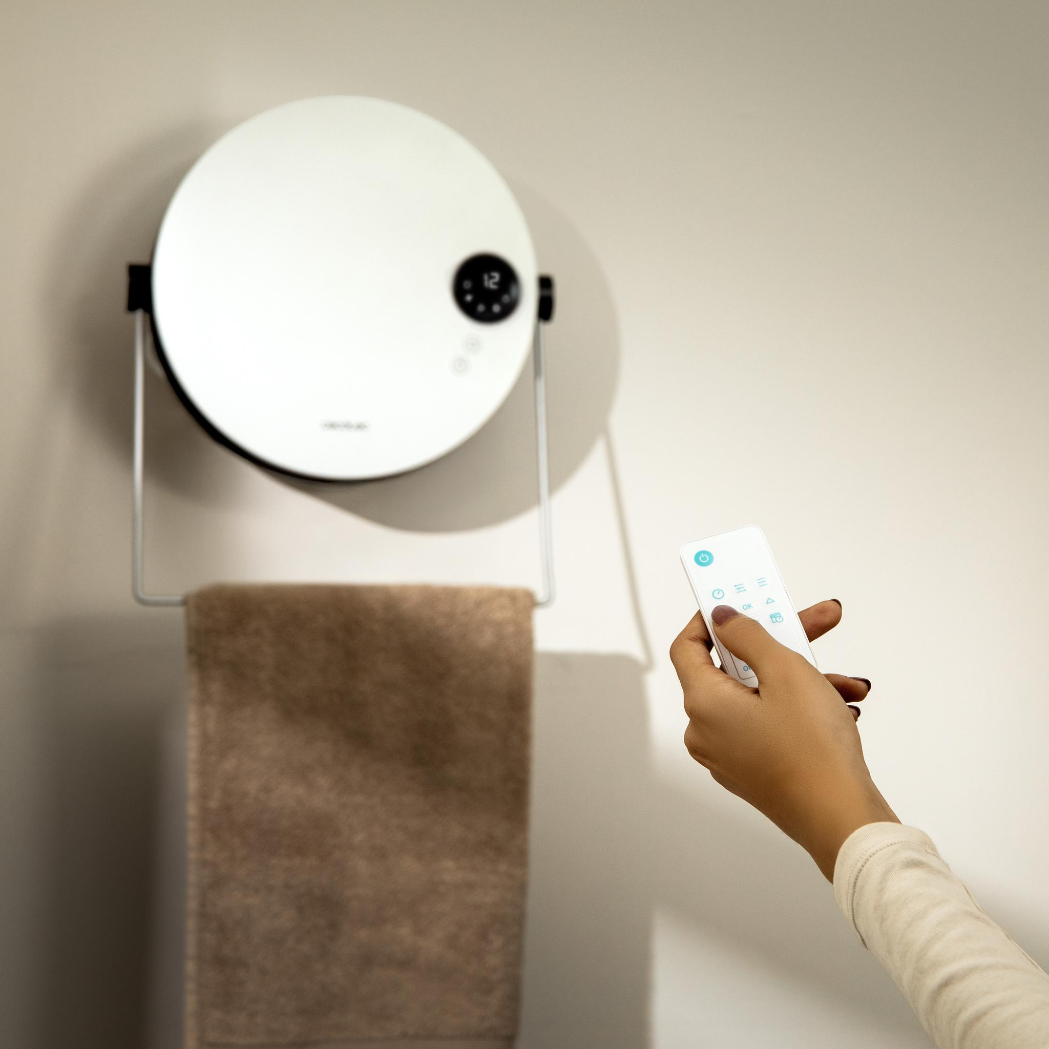 Una mano sosteniendo un mando a distancia lo apunta hacia un calefactor circular blanco en la pared con una toalla debajo.