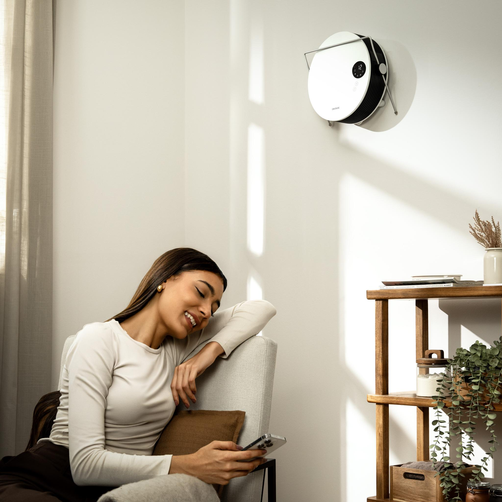 Mujer sonríe, sosteniendo teléfono, sentada en sofá. Purificador de aire en pared, estante con plantas y decoración, luz de ventana.