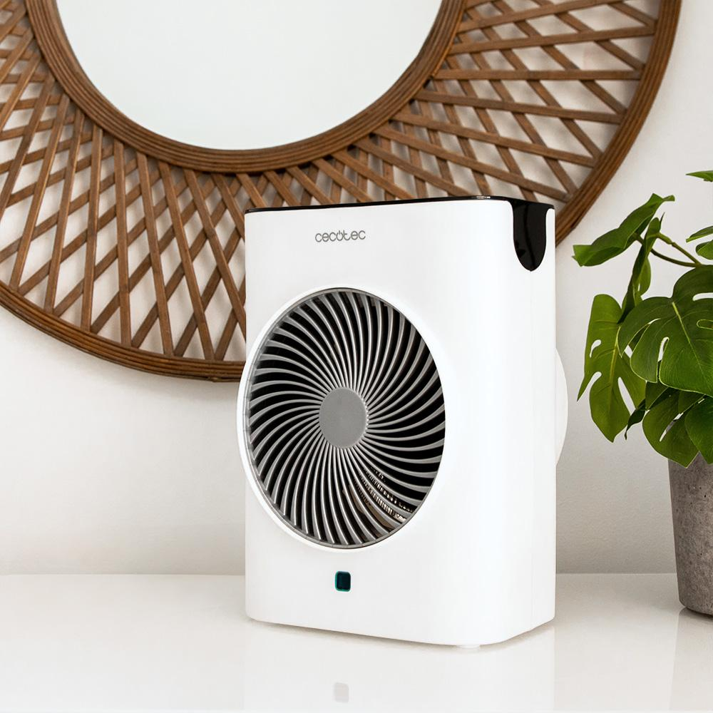 Un ventilador blanco con detalles en negro sobre una superficie blanca. Un espejo de madera y una planta al fondo.