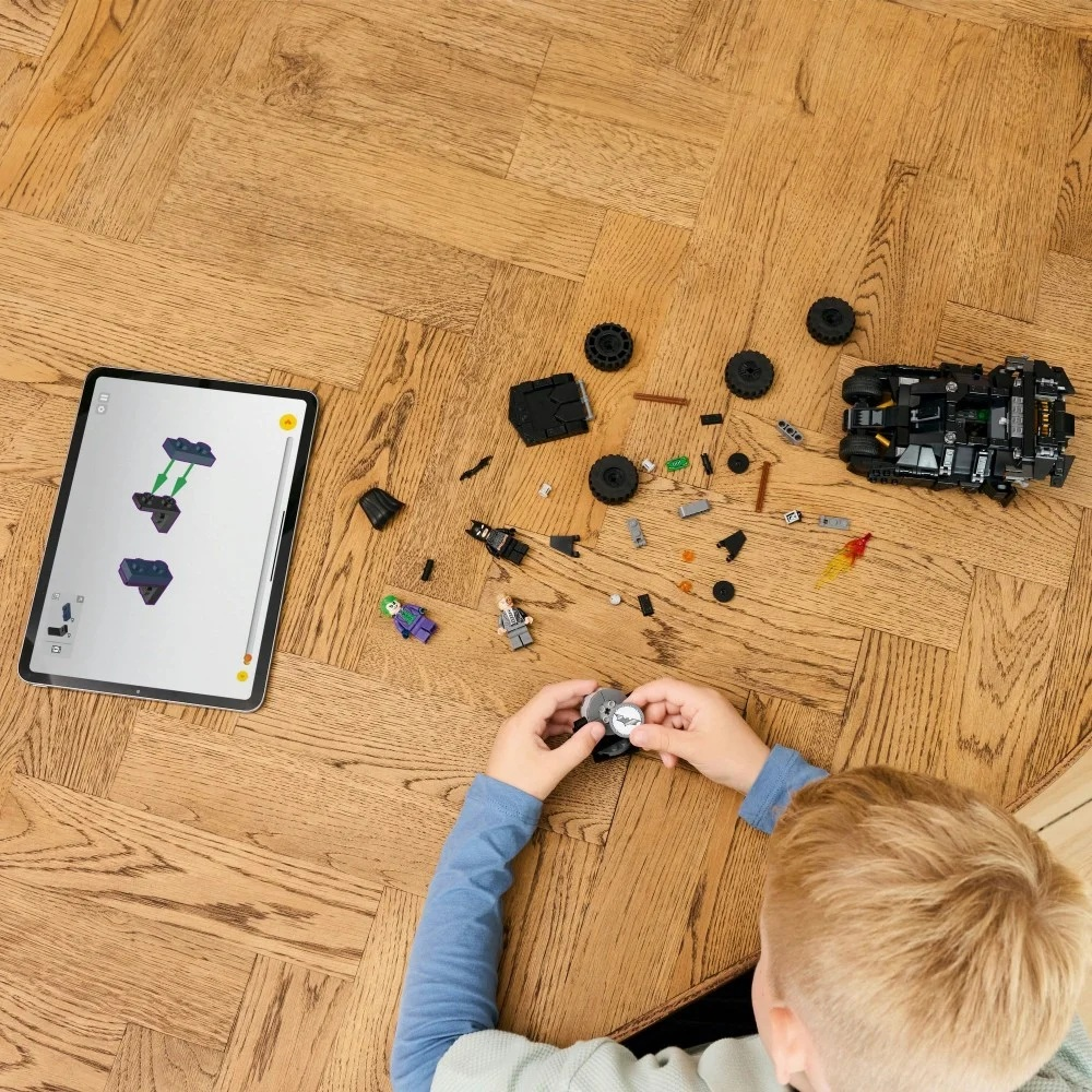 Dziecko składa zestaw Lego na drewnianej podłodze. Tablet wyświetla instrukcje. Klocki Lego i figurki są rozrzucone.