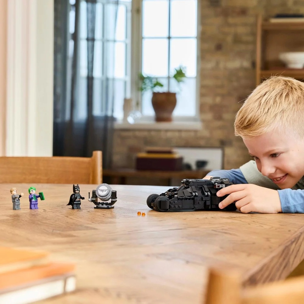 Chłopiec uśmiecha się do Batmobilu Lego. Inne figurki Lego i reflektor są na drewnianym stole. W tle okno i ceglana ściana.
