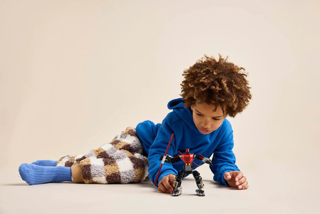 Dziecko w niebieskiej bluzie z kapturem i wzorzystych spodniach bawi się zabawką robota. Tło jest beżowe.