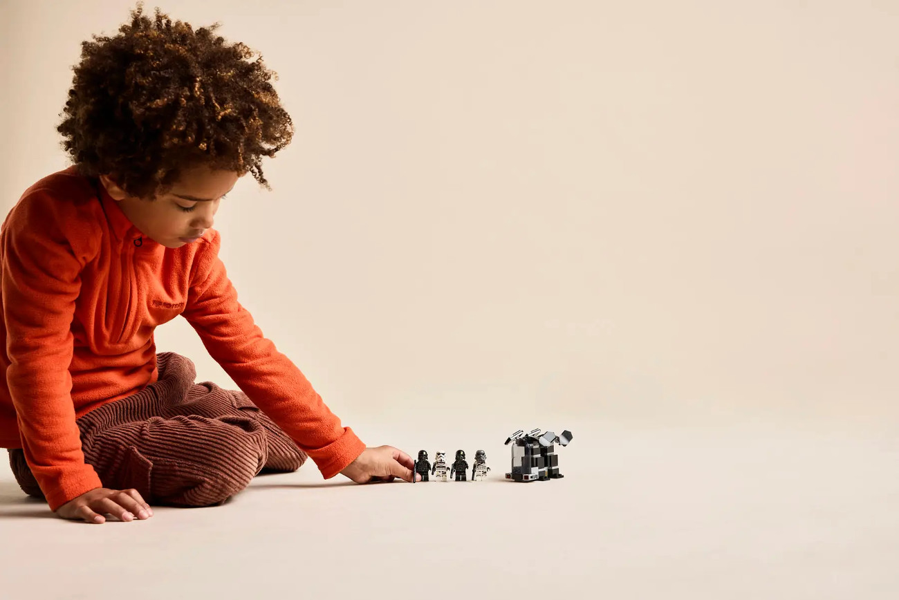 Dziecko w pomarańczowym siedzi i sięga po figurki Lego i konstrukcję. Tło jest beżowe.