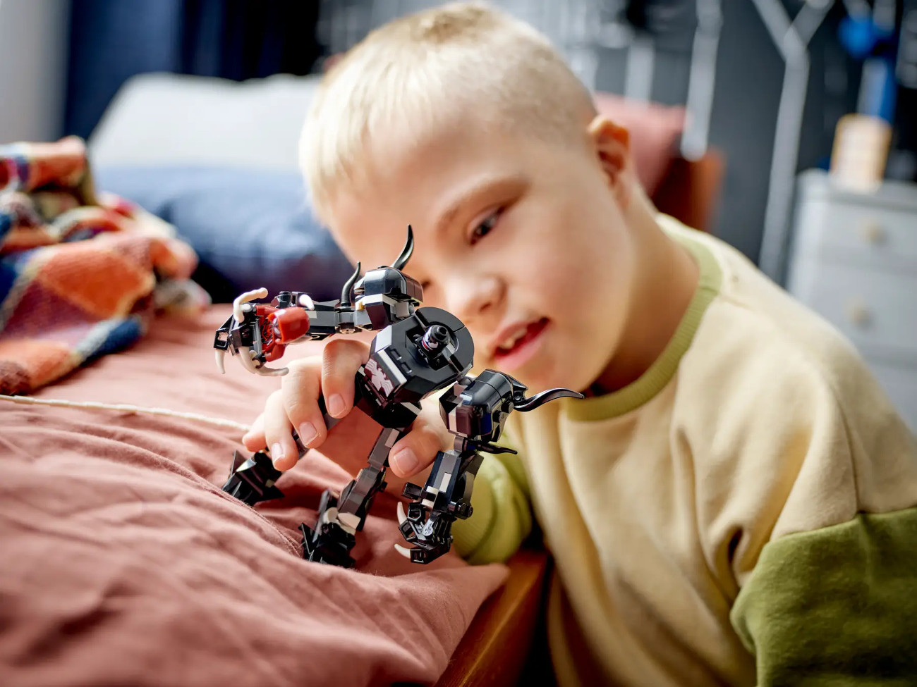 Mały chłopiec trzyma czarną zabawkę robota. Robot jest na łóżku. Chłopiec ma na sobie żółtą koszulę z zielonymi rękawami.