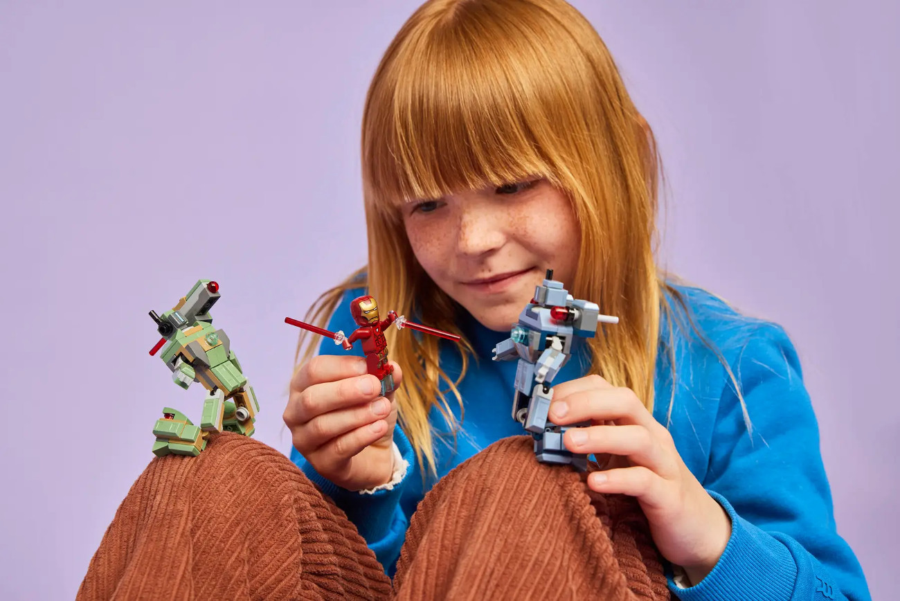 Dziewczyna z rudymi włosami trzyma Lego Iron Mana i dwie figurki robotów. Ma na sobie niebieski sweter i siedzi na brązowej tkaninie.