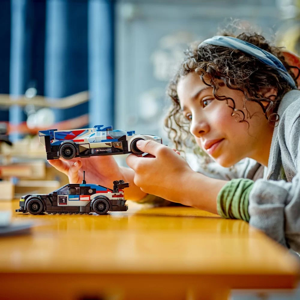 Dziewczyna trzyma dwa samochodziki wyścigowe Lego. Jeden jest biało-niebieski, drugi czarno-niebieski, oba na drewnianym stole.