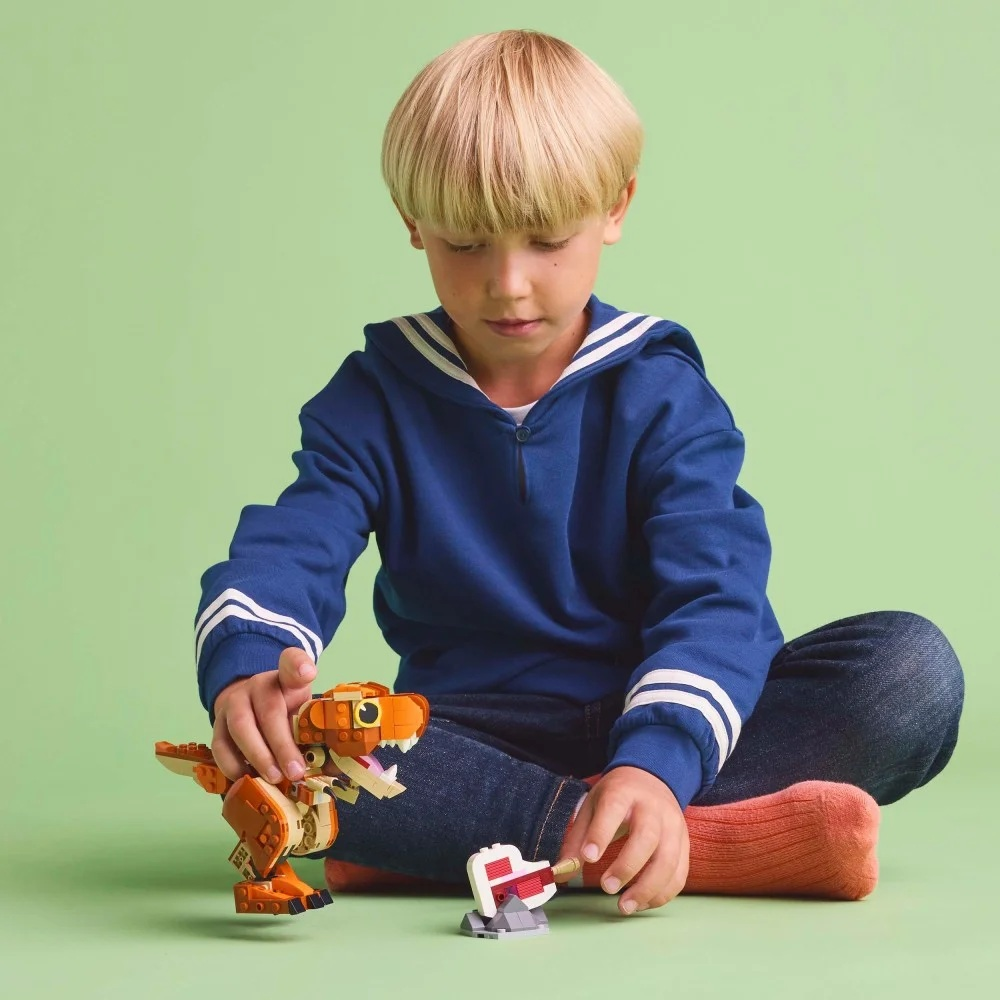 Chłopiec z blond włosami siedzi na podłodze, budując dinozaura z Lego. Ma na sobie niebieski sweter i dżinsy. Zielone tło.