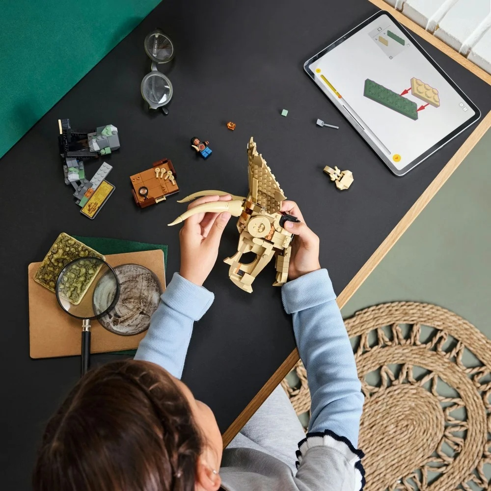 Dziecko składa czaszkę dinozaura Lego na czarnym stole z otwartym tabletem pokazującym instrukcje. W pobliżu znajdują się inne klocki Lego, okulary i szkło powiększające.