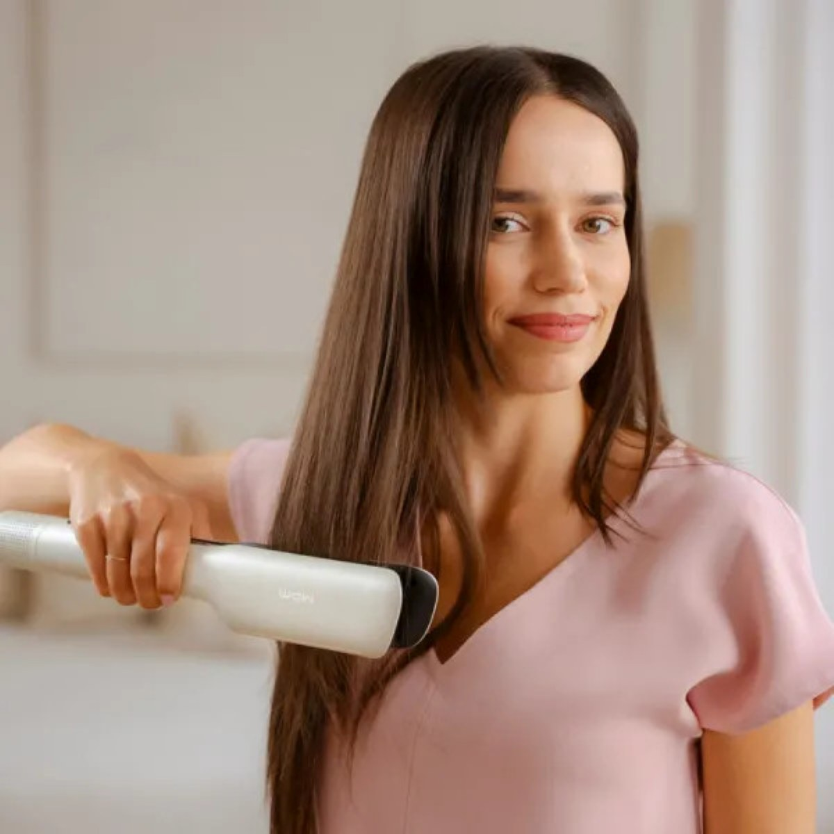 Kobieta z długimi brązowymi włosami używa prostownicy. Ma na sobie różowy top. Tło to rozmyty pokój.