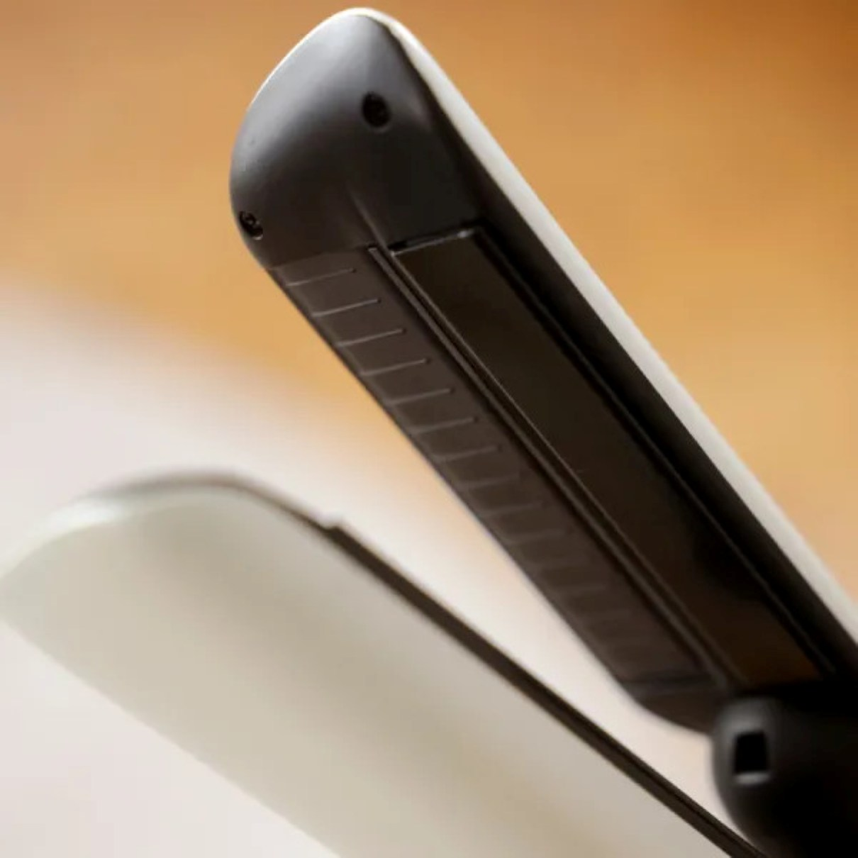 Zbliżenie czarno-białej prostownicy do włosów. Tło jest rozmyte, ukazując odcienie pomarańczu i brązu.