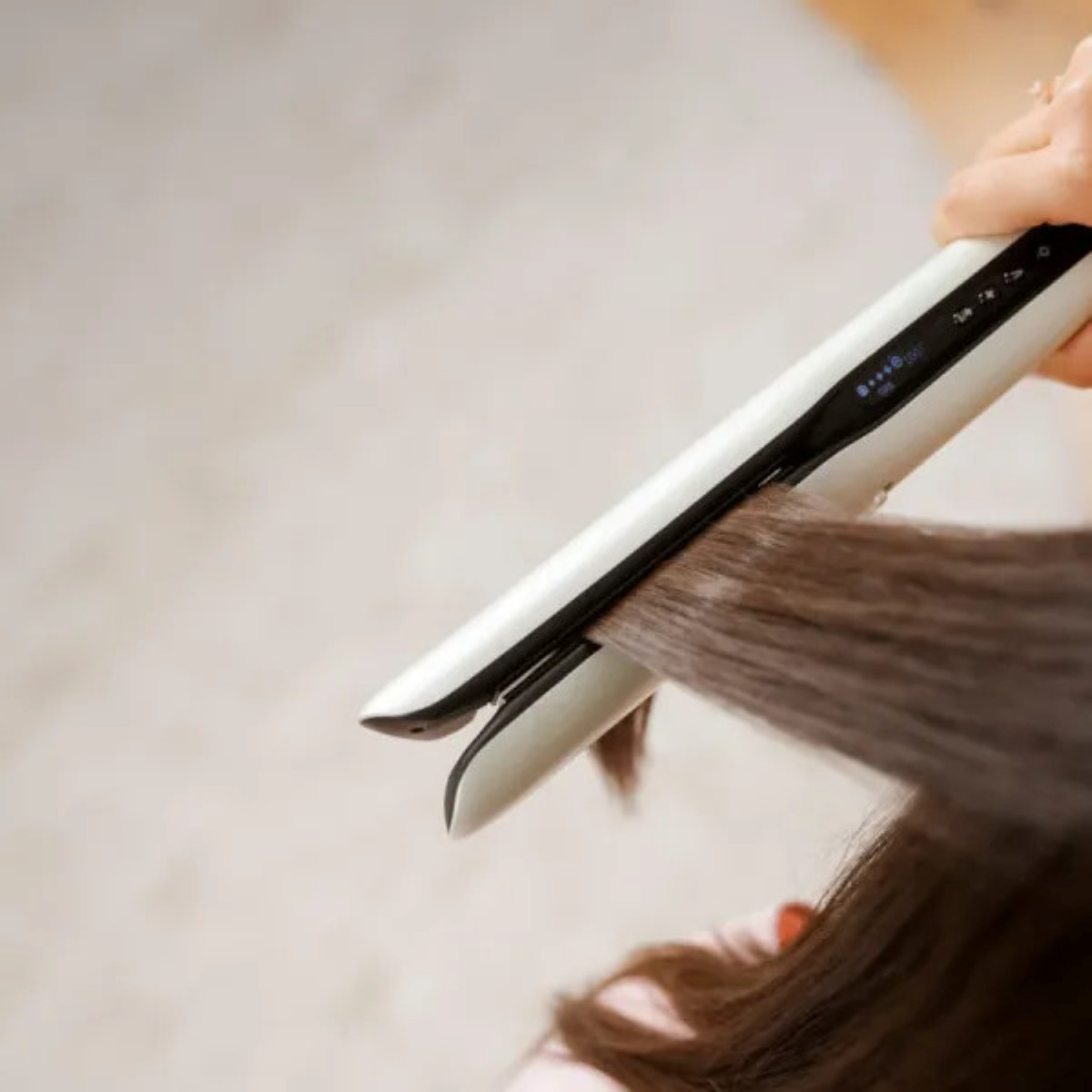 Osoba używa białej prostownicy do brązowych włosów. Ręka trzyma urządzenie, z jasnym tłem.