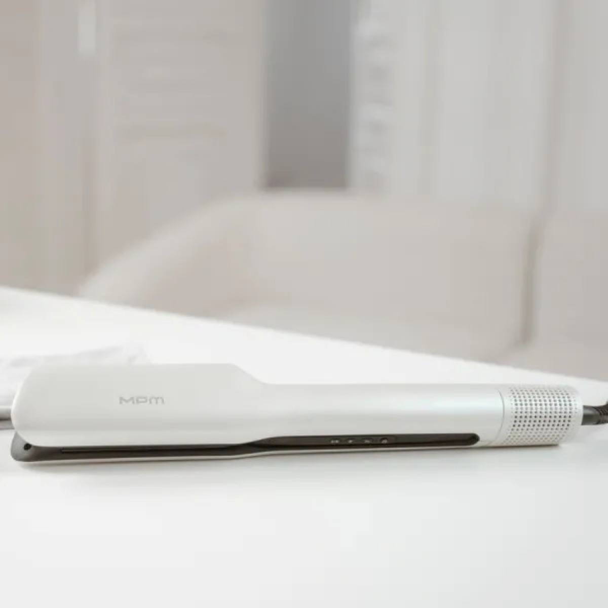 Biała prostownica do włosów z napisem 'MDM' leży na białym stole. Tło to rozmyte białe pomieszczenie.