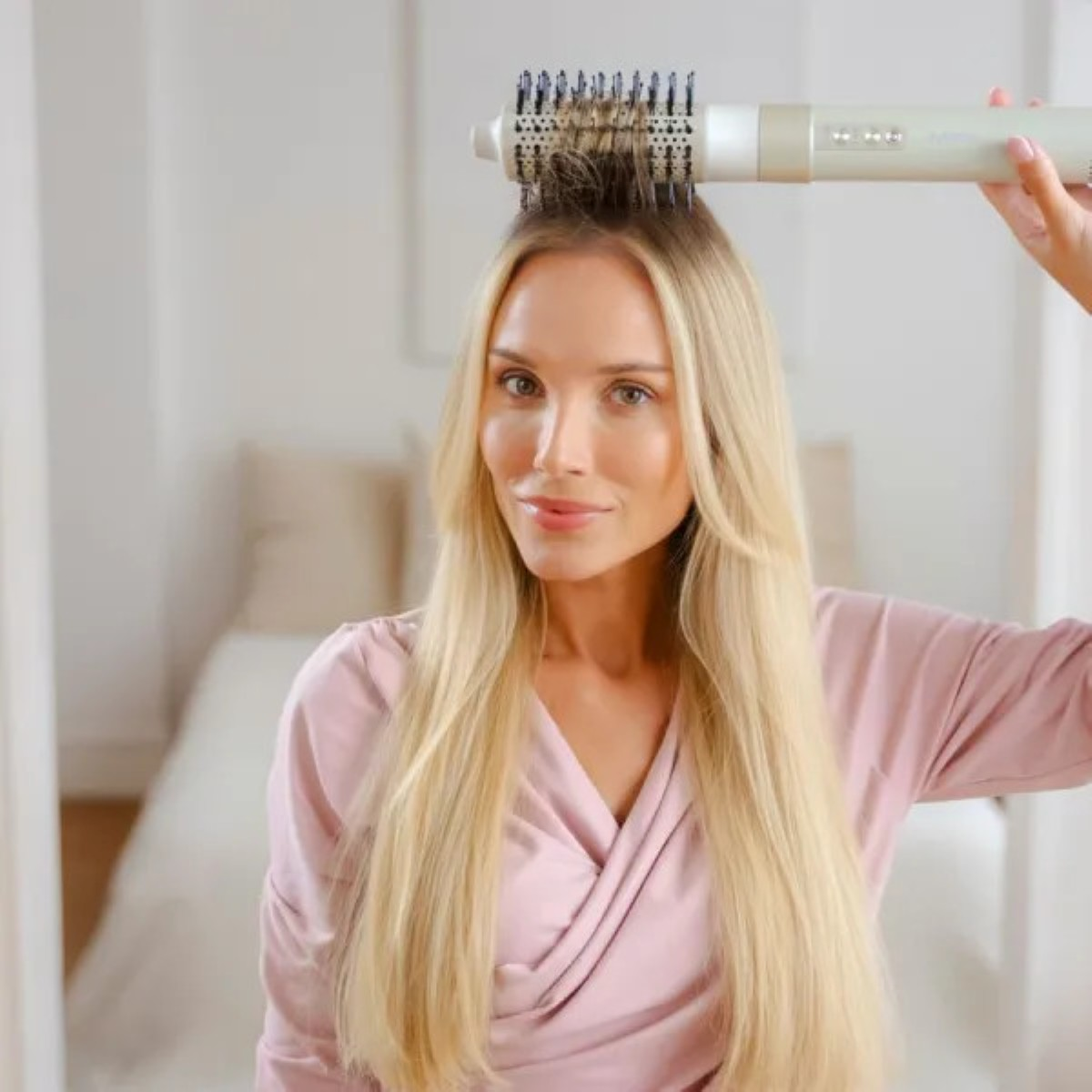 Blondynka używa narzędzia do stylizacji włosów. Ma na sobie różowy top. Tło obejmuje łóżko i neutralną dekorację.