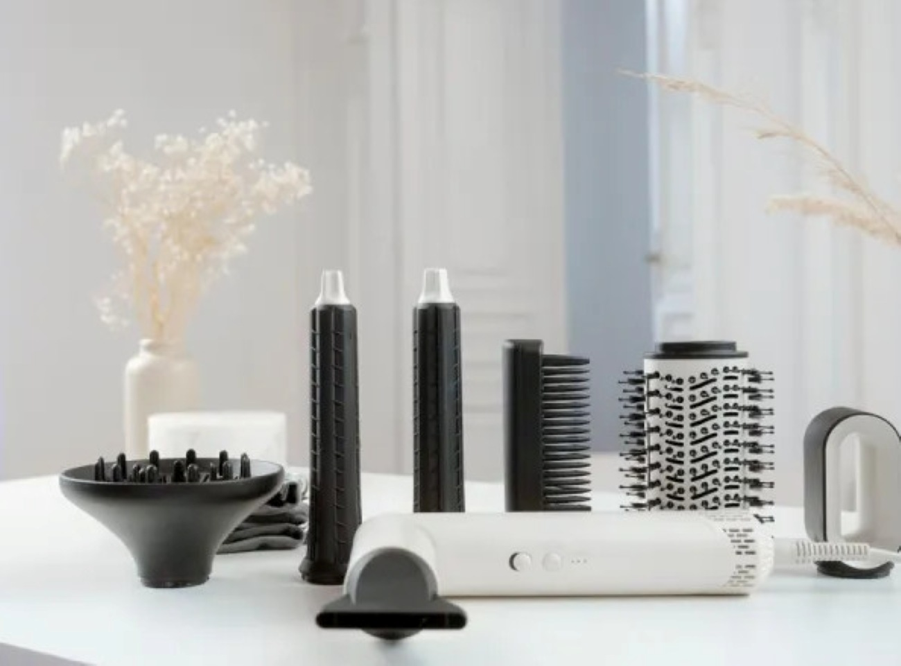 Narzędzia do stylizacji włosów: suszarka, czarne nasadki, biały uchwyt i szczotki. Suszone kwiaty w wazonie, wszystko na białej powierzchni.