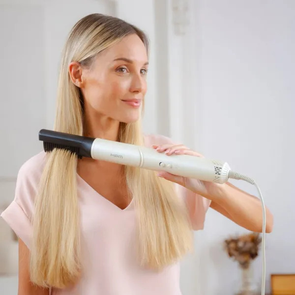 Kobieta szczotkuje swoje długie blond włosy biało-czarnym narzędziem do stylizacji włosów, uśmiechając się w jasnym pokoju.
