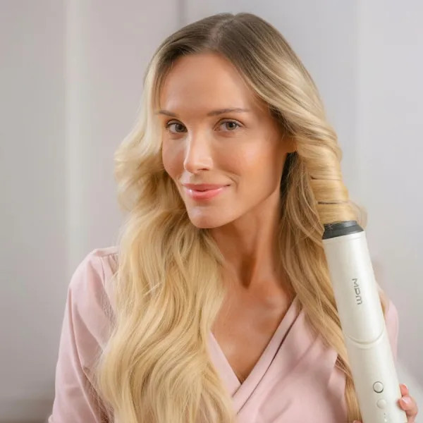 Kobieta z blond włosami kręci włosy białą lokówką. Ma na sobie różowy szlafrok i uśmiecha się do kamery.