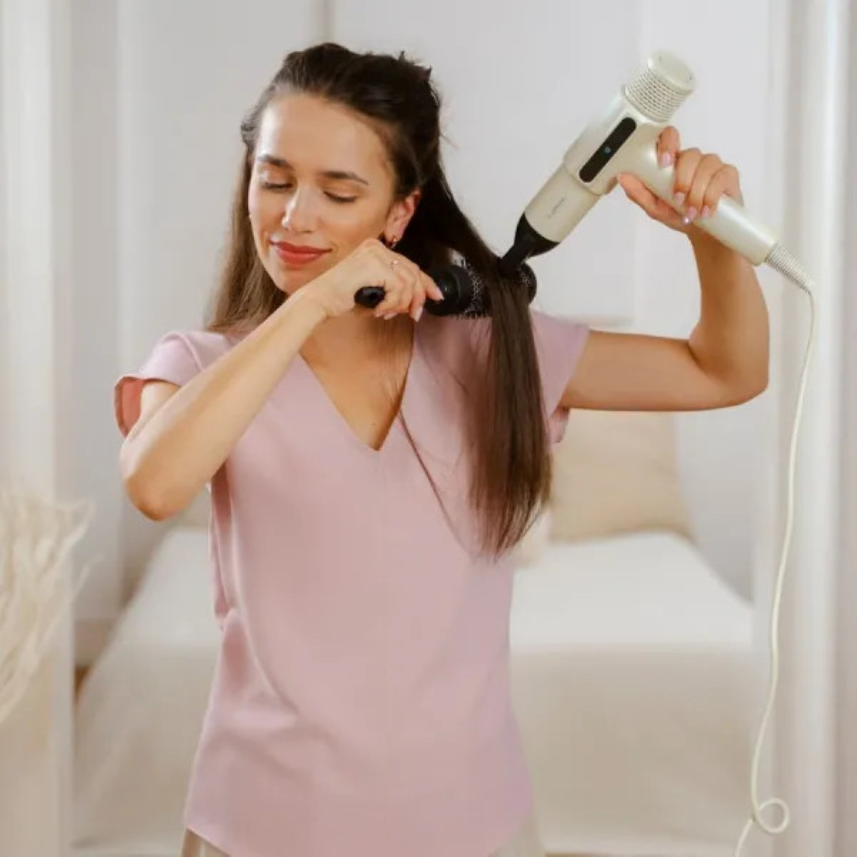 Kobieta w różowej koszuli suszy włosy. Trzyma szczotkę i suszarkę. Tło to sypialnia.