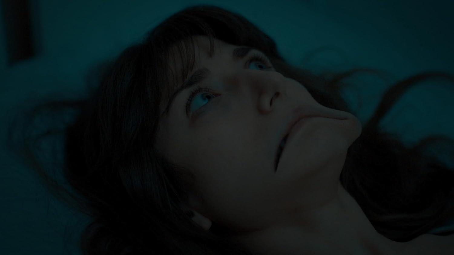 Das Gesicht einer Frau, die nach oben schaut. Ihre Augen sind blau. Dunkle Haare und Haut. Der Hintergrund ist ein schwach, teal-farbener Raum.