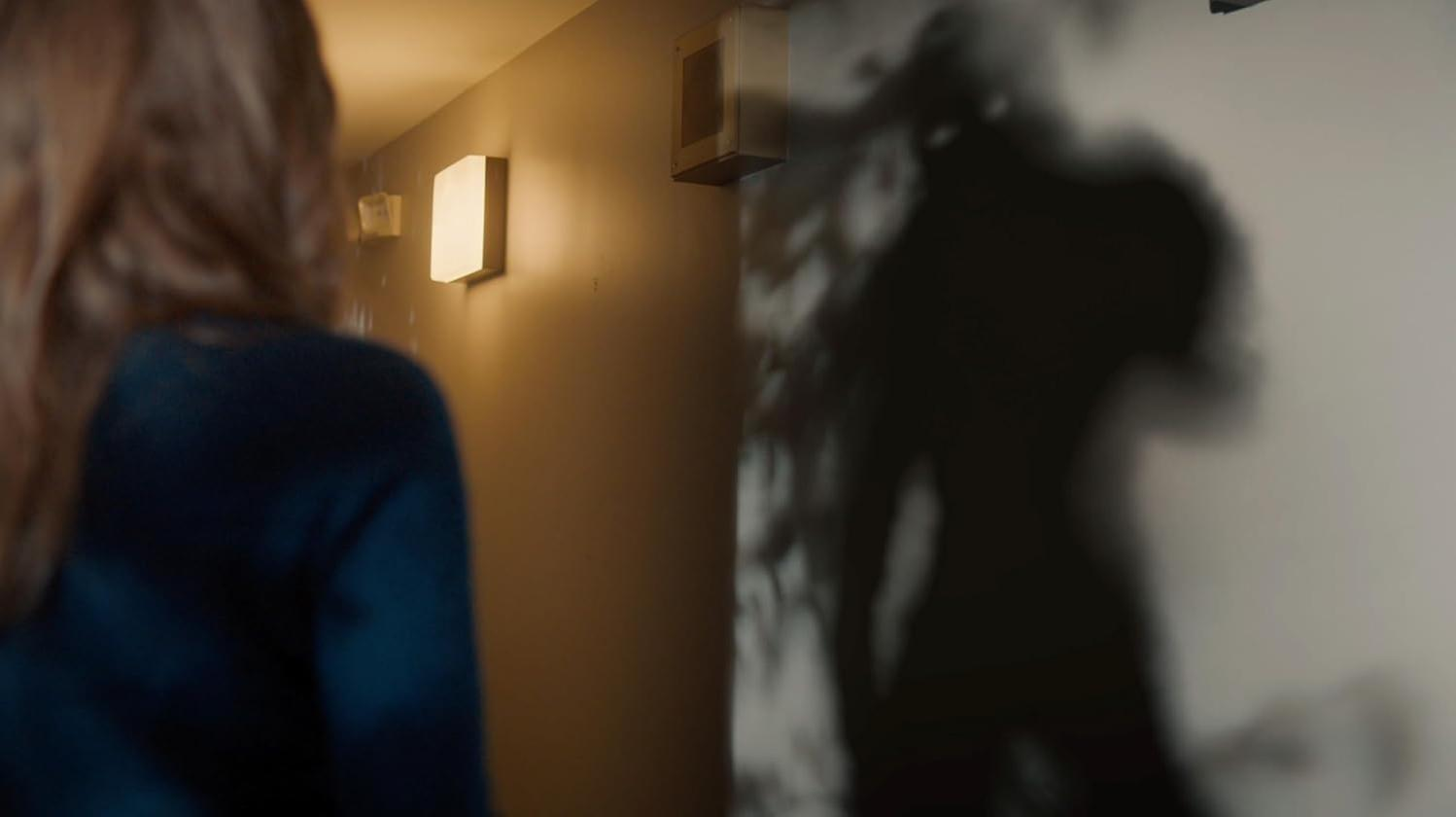 Eine Frau geht einen Flur entlang. Ein großer Schatten wird auf die Wand geworfen. Lichter sind an der Wand.