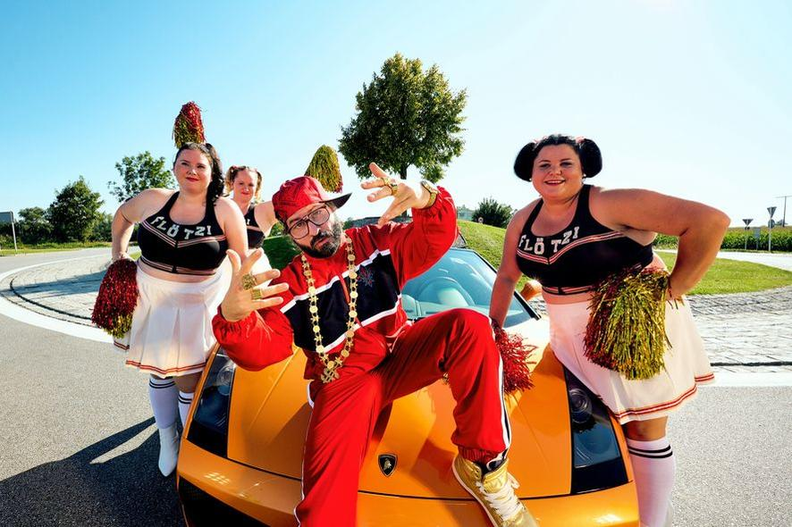 Ein Mann in rotem Trainingsanzug posiert auf einem orangenen Auto mit Cheerleadern. Sie stehen auf einer Straße mit Bäumen und blauem Himmel.