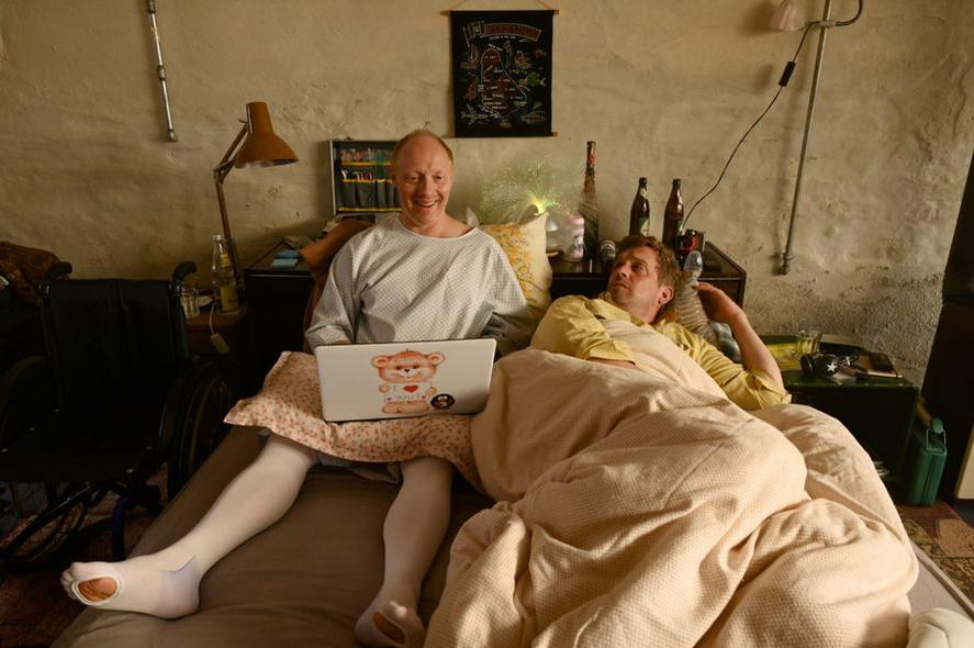 Zwei Männer in einem Zimmer. Einer benutzt einen Laptop mit Teddybär-Aufkleber. Der andere liegt im Bett. Flaschen und ein Poster sind sichtbar.