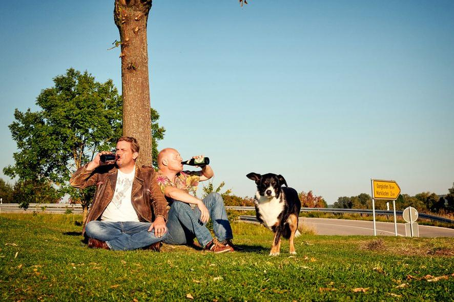 Zwei Männer trinken Bier unter einem Baum mit einem Hund. Im Hintergrund ein Straßenschild, eine Straße und ein klarer blauer Himmel.