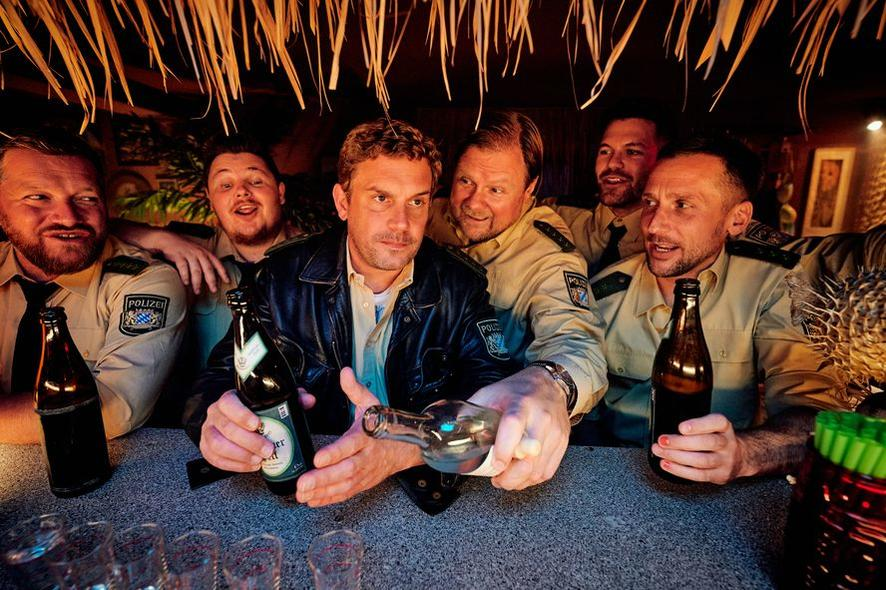Eine Gruppe von Polizisten in einer Bar, die Flaschen halten. Einer gießt aus einer Flasche. Die Szene hat eine warme, einladende Atmosphäre.