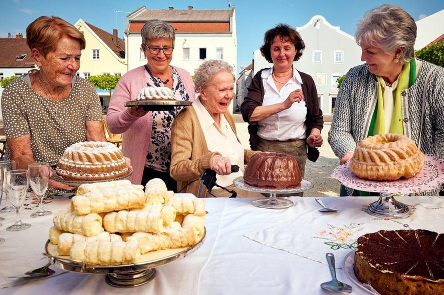 Fünf Frauen mit Kuchen, Gebäck und einer Frau im Rollstuhl an einem Tisch. Gebäude im Hintergrund. Die Stimmung ist festlich.
