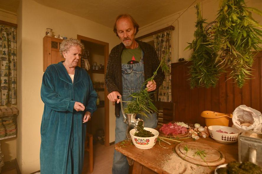 Eine ältere Frau in einem tealfarbenen Bademantel steht neben einem Mann, der Kräuter mahlt. Kräuter hängen von der Decke, Lebensmittel auf dem Tisch.