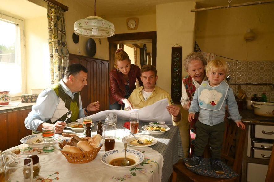 Familie am Tisch, betrachtet ein Papier. Ein Kind steht auf einem Stuhl. Essen und Getränke auf dem Tisch. Warm und einladend.