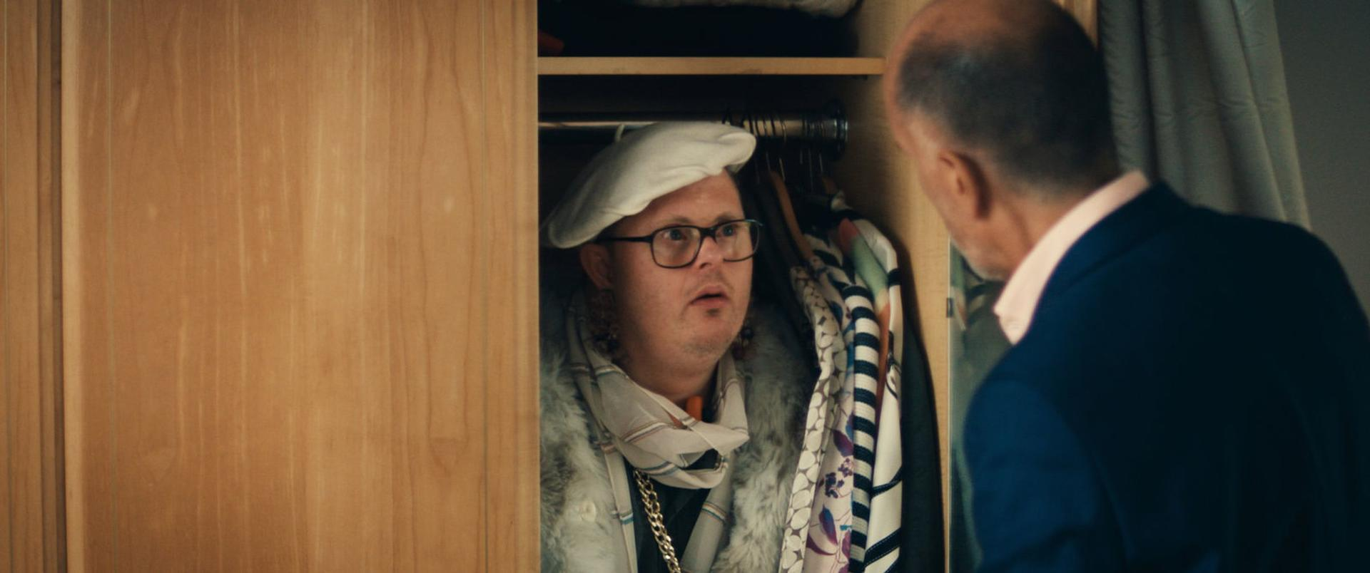 Mann mit weißer Baskenmütze und Brille im Schrank, sieht überrascht aus. Ein anderer Mann im Anzug schaut ihn an.