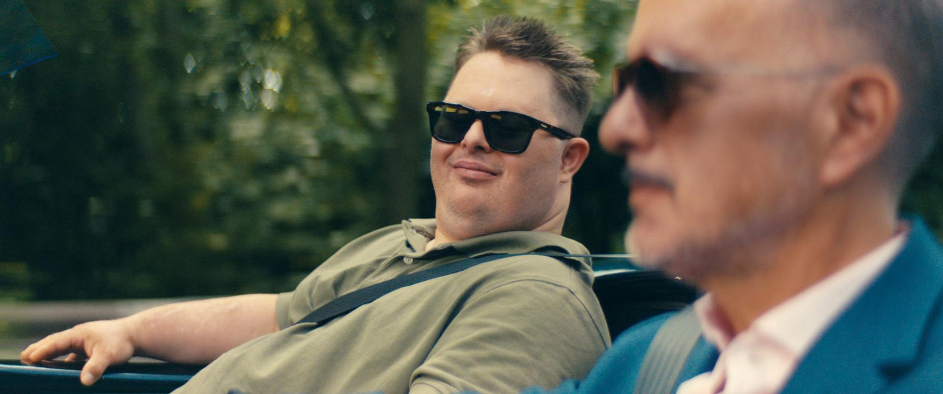 Zwei Männer in einem Auto. Einer trägt eine Sonnenbrille und ein grünes Hemd. Der andere ist verschwommen, trägt eine blaue Jacke.