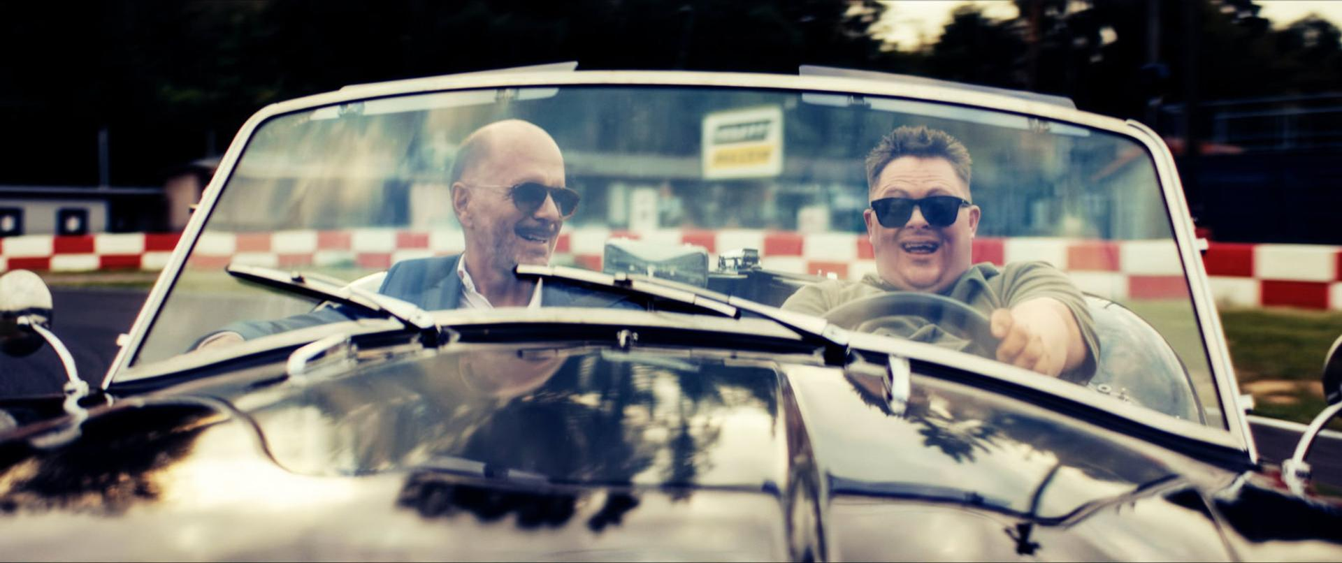Zwei Männer mit Sonnenbrille fahren einen Oldtimer. Einer fährt, der andere lächelt. Rennstrecke im Hintergrund.