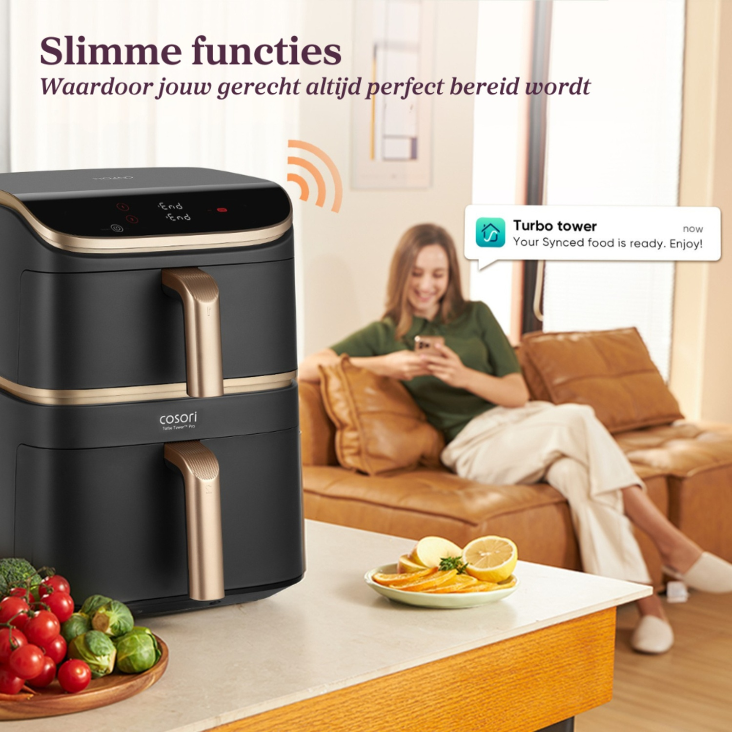 An air fryer with a woman on a couch. The fryer is black with gold accents and vegetables are on the table.