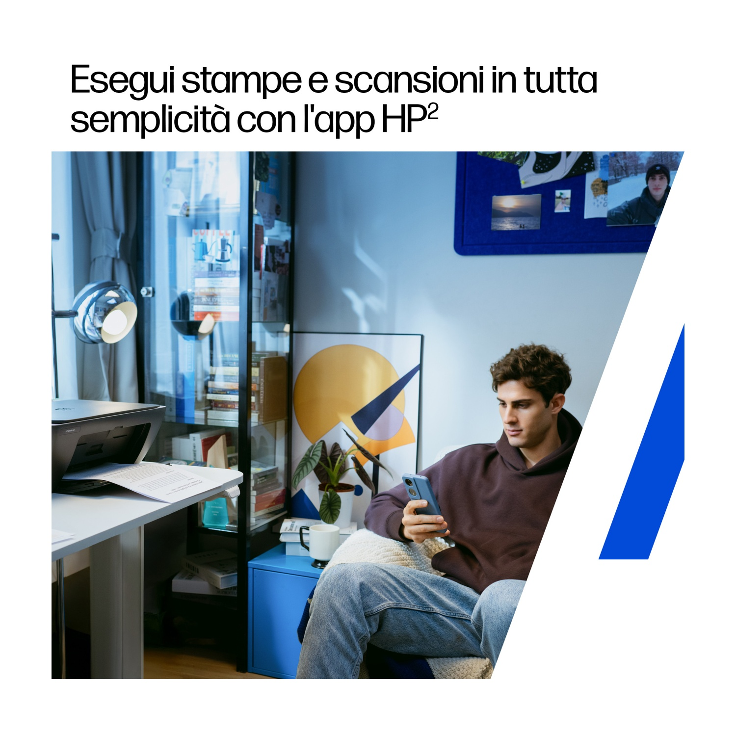 Un uomo con una felpa con cappuccio bordeaux usa il telefono. Una stampante e una scrivania sono in primo piano. Un mobile blu.