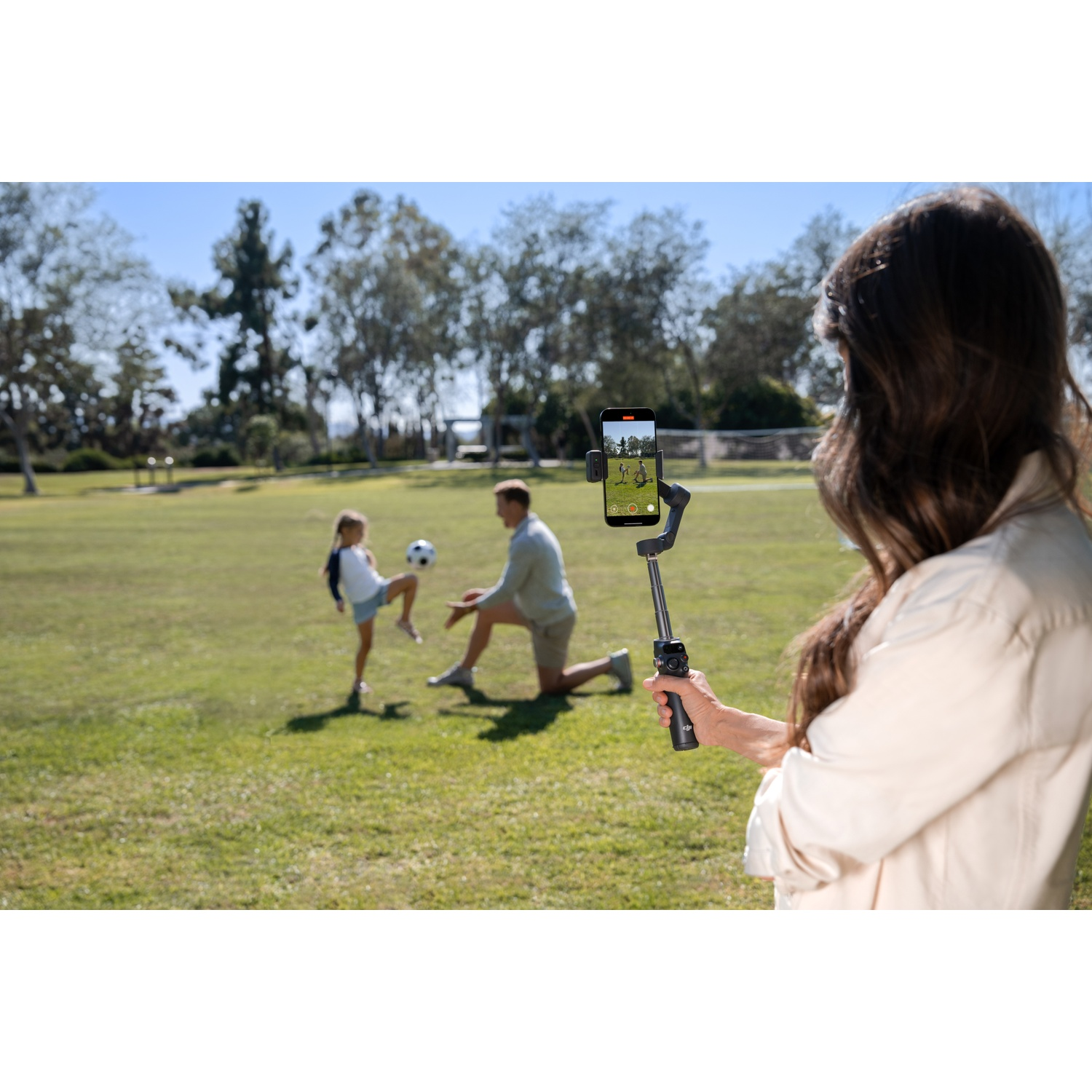 A woman films a family playing soccer on a phone attached to a selfie stick. A girl kicks the ball, a man kneels.