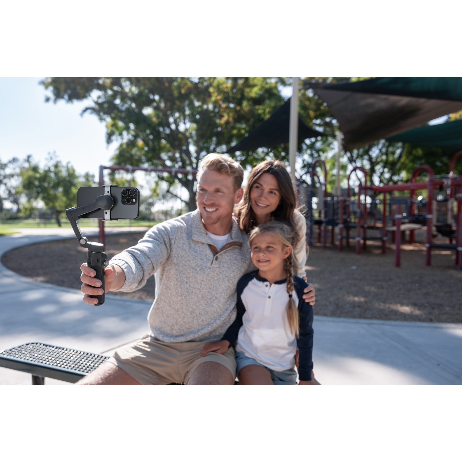 Family taking a selfie with a phone on a gimbal at a park. Man holds the gimbal, with woman and girl beside him.