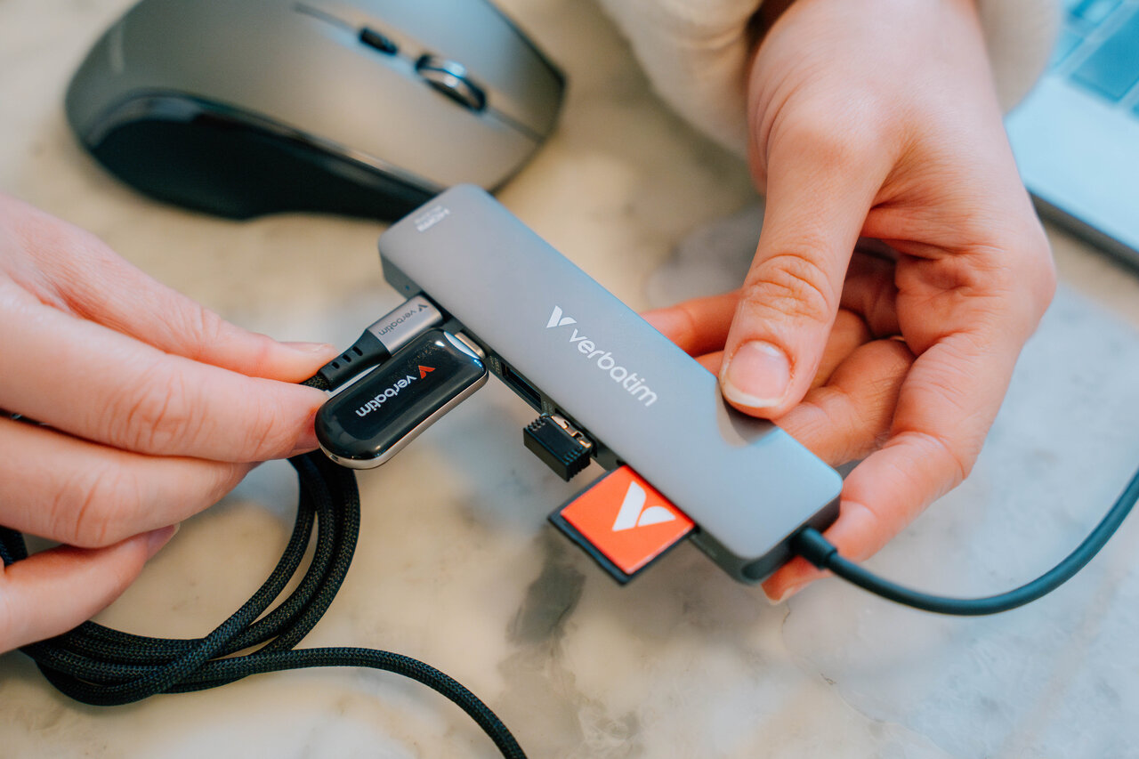 Eller, kablolar ve bir hafıza kartı bağlayan bir Verbatim USB hub'ı tutuyor. Arka planda bir fare görünüyor.