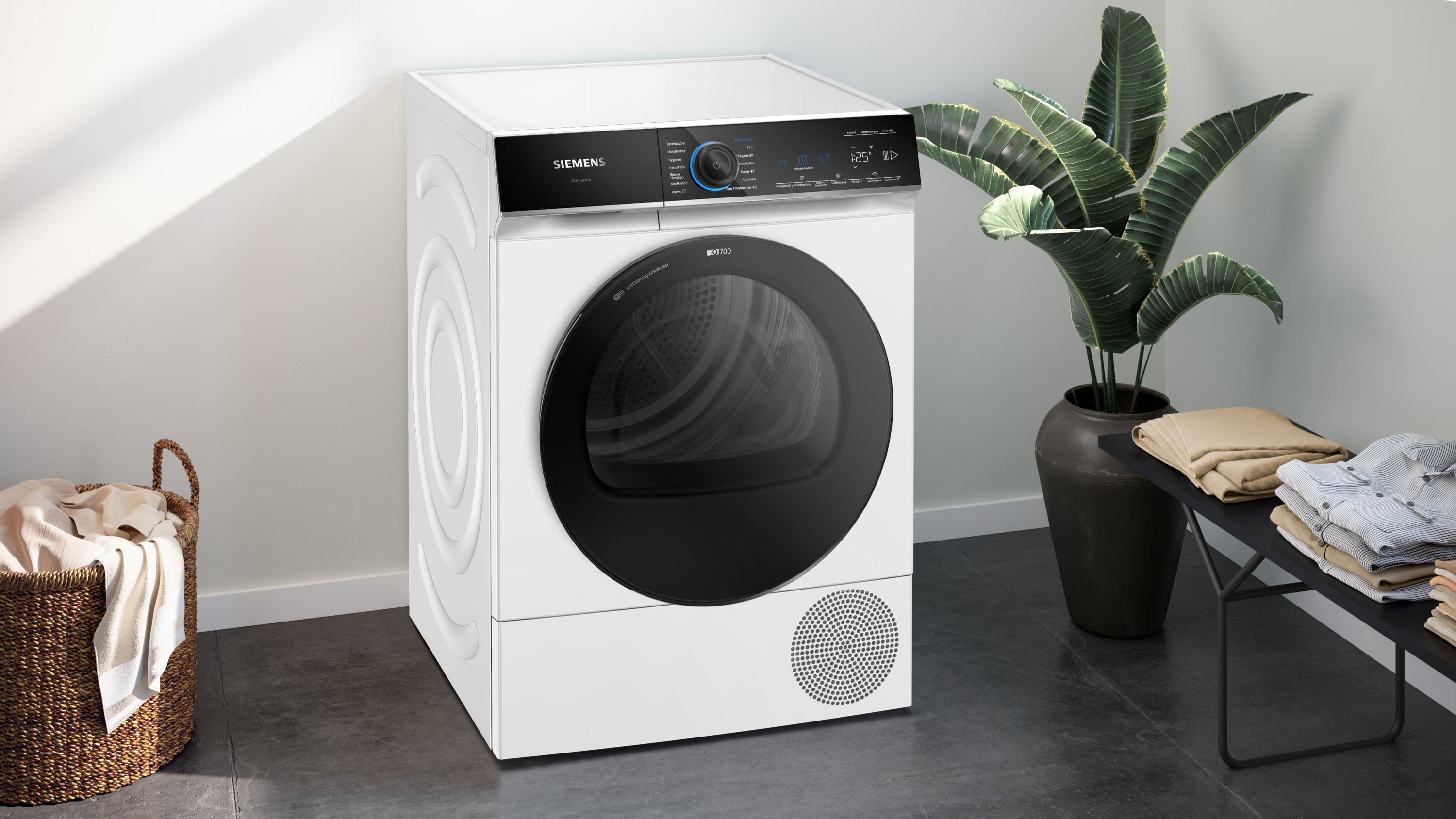 Ein weißer Siemens-Trockner mit schwarzer Tür, neben einer Pflanze und einem Korb mit Kleidung.