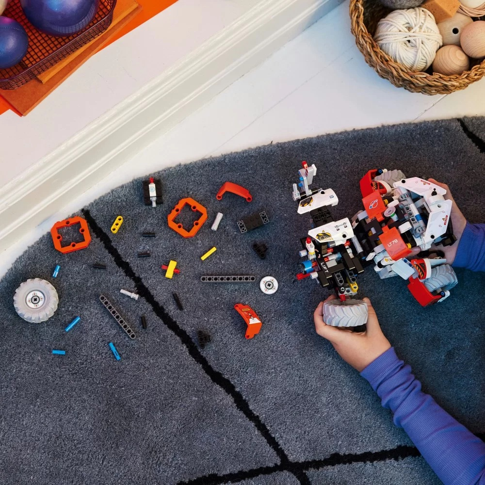 Dziecko składa model Lego na szarej wykładzinie. Różne klocki Lego są rozrzucone, a częściowo zbudowany model jest w centrum uwagi.