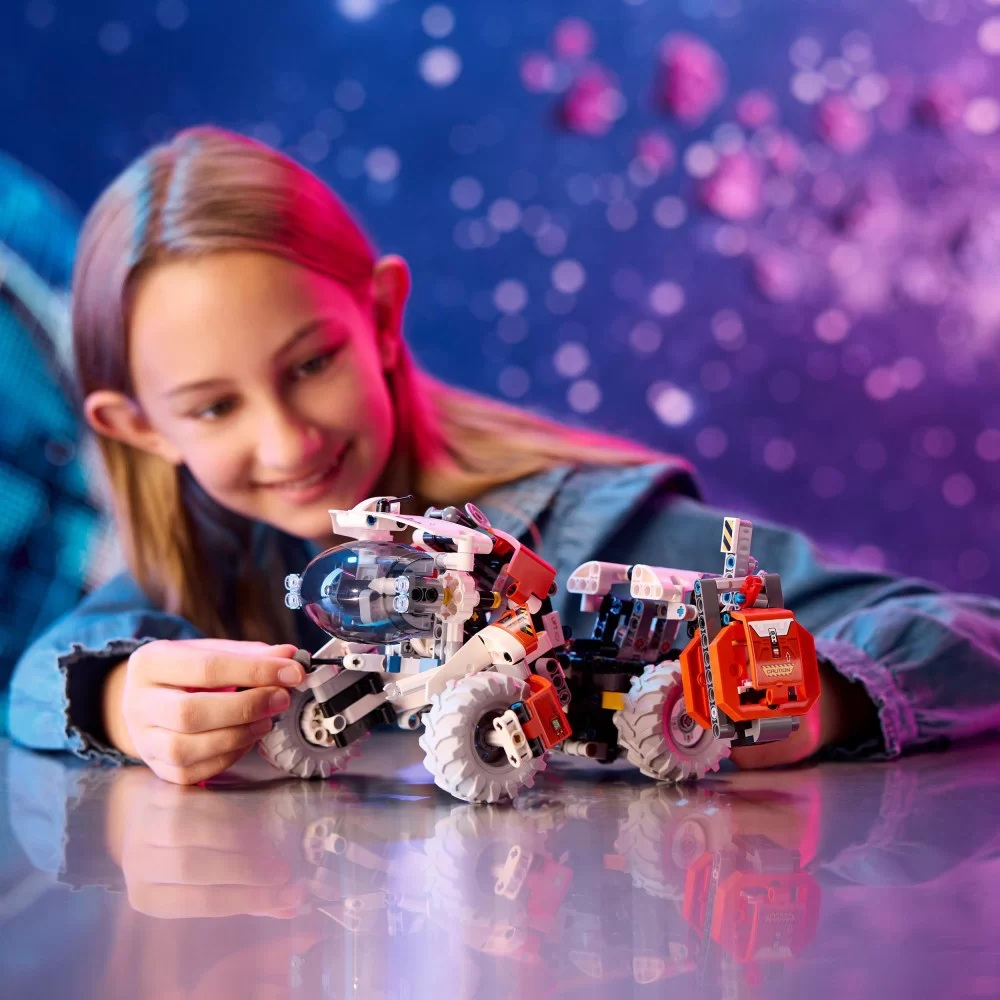 Dziewczyna uśmiecha się do zabawek. Jedna jest biało-czerwona, druga pomarańczowa. Tło to scena podobna do galaktyki.