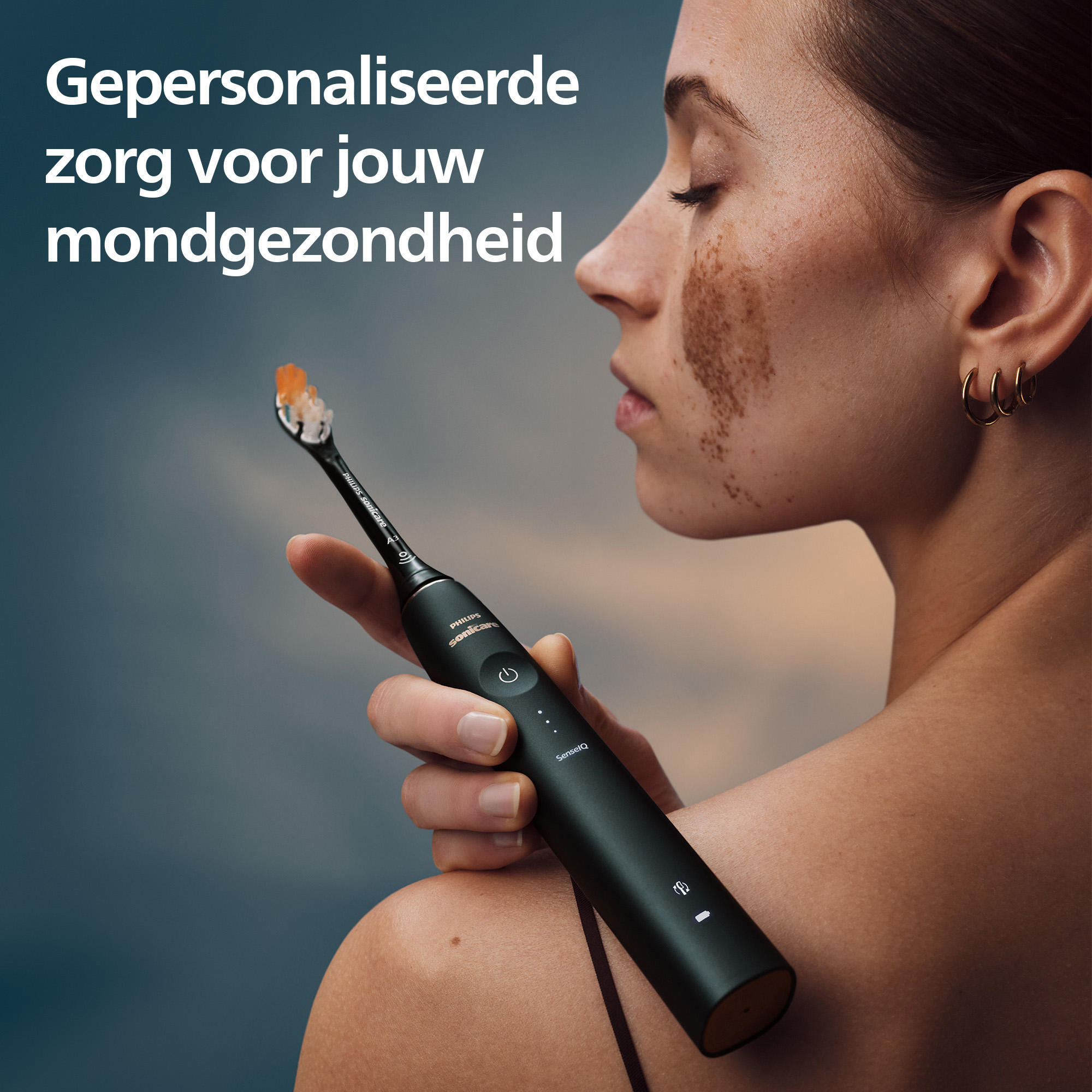 Woman holding a black electric toothbrush. She has freckles and gold earrings. The background is a gradient of blue and orange.