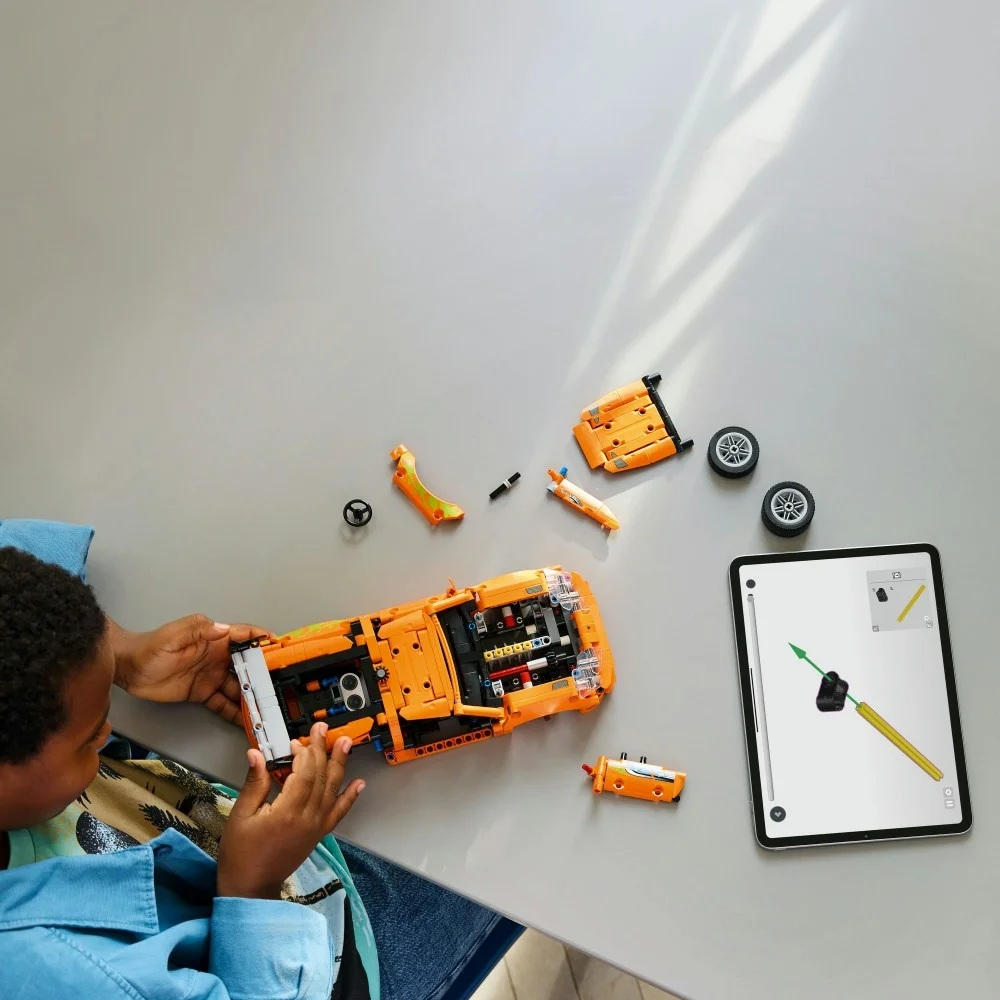 Dziecko składa pomarańczowy samochód Lego na stole. W pobliżu znajdują się części i tablet z instrukcjami.